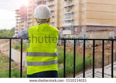 Woman Builder in a white helmet and yellow vest. Middle-aged woman working on a construction site. Her back is to the camera