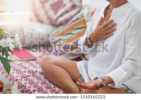 Self-Healing Heart Chakra Meditation. Woman sitting in a lotus position with right hand on heart chakra and left palm open in a receiving gesture. Self-Care Practice at Home Royalty-Free Stock Photo #1510165223