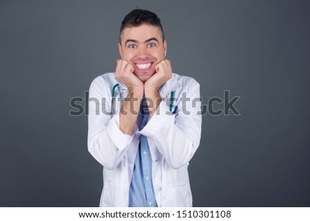 Dreamy young European doctor man keeps hands pressed together under chin, looks with happy expression, has toothy smile, isolated over gray background