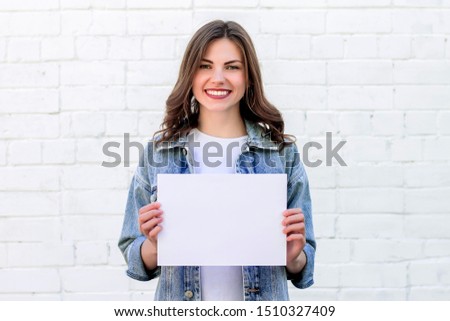 Girl student smiling and holding a white sheet of paper on a background of a white brick wall