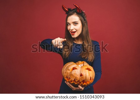 Woman dressing in black devil Halloween costume with witch's headband for a themed party. She hold pumpkin for a 'trick or treat' October festival. Isolated on red background. Witchcraft concept.