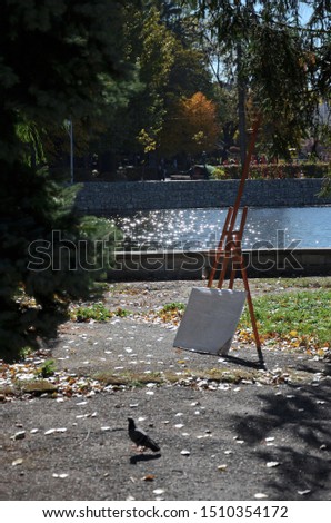 In the park near the lake there is an easel with white canvas and a dove in the foreground