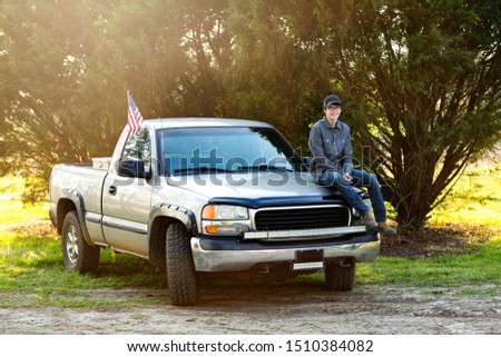 happy teenage boy from the south sitting on the front of his pickup truck outside in a rural area. 