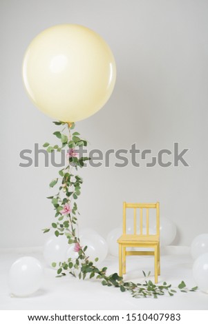 Beautiful decor for a children's photo shoot. A large yellow balloon with a garland of real flowers in the air. White balloons and children’s yellow wooden chair on a white background. Studio light.