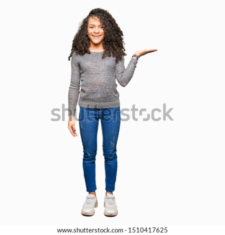 Young beautiful woman with curly hair wearing grey sweater smiling cheerful presenting and pointing with palm of hand looking at the camera.