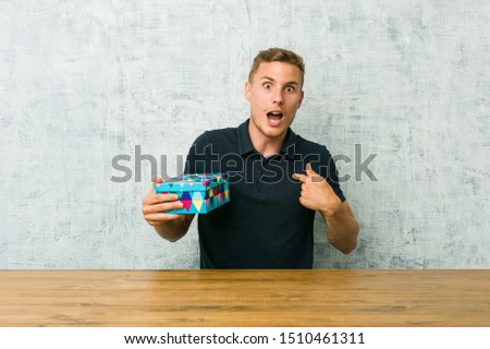 Young caucasian man holding a gift box on a table surprised pointing with finger, smiling broadly.
