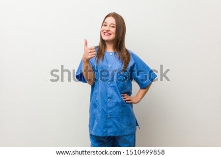 Young nurse woman against a white wall smiling and raising thumb up