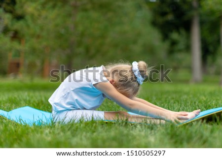 A little disgruntle blue-eyed, curly blonde girl in a black and red gymnastic suit is doing stretching on the blue yoga carpet in the park