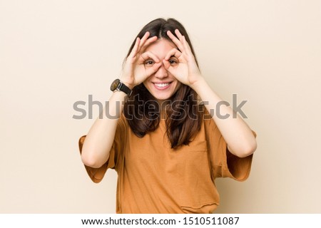 Young brunette woman against a beige background showing okay sign over eyes