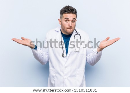 Young handsome doctor man confused and doubtful shrugging shoulders to hold a copy space.