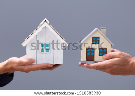 Close-up Of Couple's Hand Holding Different Miniature Houses Against Gray Background Royalty-Free Stock Photo #1510583930