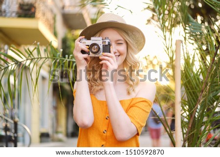 Pretty smiling blond girl in hat happily taking photo on retro camera on city street