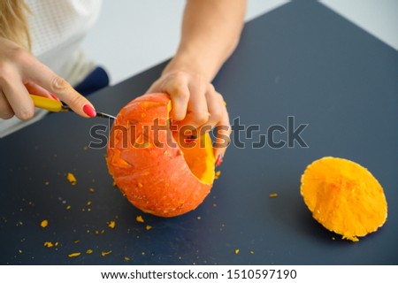 A woman cuts out a face with a stationery knife on a pumpkin for Halloween lamps on a black table. Close-up of female hands making jack-o'-lantern on the eve of all saints. Front view