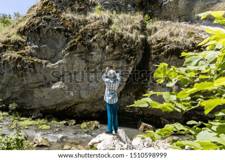 A teenager in turquoise trousers stands on a stone near the river and take a photo the rock with moss on a mobile phone in the bright sunlight. Concept young photographer interested in nature.