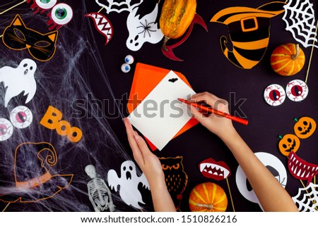 Female hands are writing greeting card on black background. Little pumpkins, photo props, masks, decor for celebration are scattered on canvas. Party accessories. Happy halloween concept.