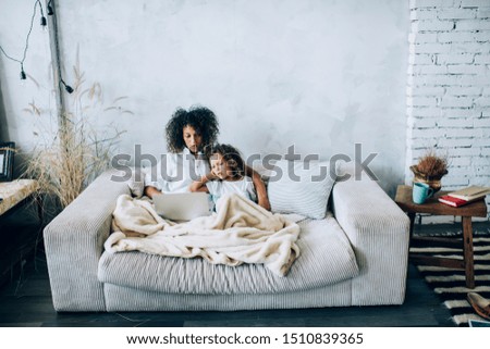 Little cute girl with beautiful mother sitting under plaid on comfy sofa at home and using laptop together in leisure