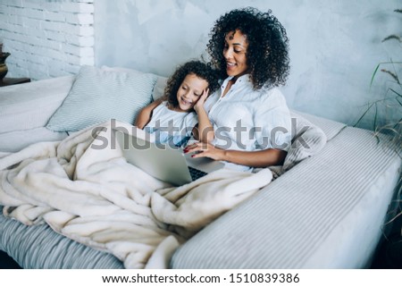 Charming happy little girl and mother with curls sitting on sofa covered with plaid and having fun together while using laptop