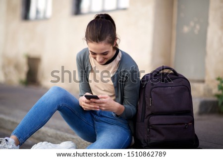 Portrait of happy female student sitting outside and looking at mobile phone 