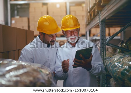 Two hardworking warehouse workers standing in warehouse and checking inventory. Older one holding tablet while younger one looking at it. Royalty-Free Stock Photo #1510878104