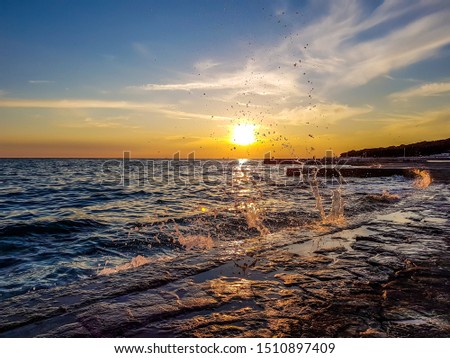 Romantic sunset by a stony harbor promenade. The sun sets over the horizon. The sunbeams reflecting in the calm sea. Stony shore is washed by waves, splashing high up. Sky is turned yellow and orange