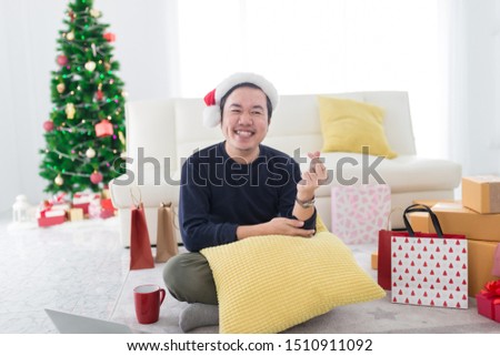 smart asian man siiting on floor, he feeling happy in celebration time, he show mini heart sign with hand, his room decorating with gift box and Christmas tree, happy new years