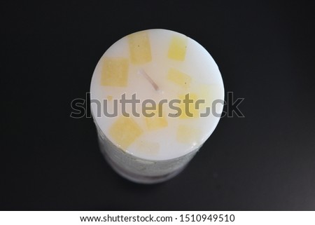 White, with yellow squares, a round, long, thick candle with a vanilla aroma is located on a black glossy surface.