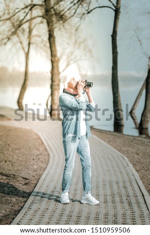 Taking pictures in the park. Stylish vigorous septuagenarian woman in denim blue clothes and short grey hair taking pictures in the park