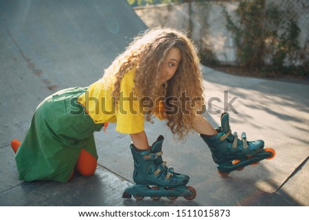 Young woman girl in green and yellow clothes and orange stockings with curly hairstyle with rollers on hand. Roller skating female in skate park.