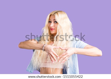 Tender romantic blonde young woman making heart shape by hand over chest, showing love sign, expressing sympathy, affection, dressed in summer clothes isolated on violet background.