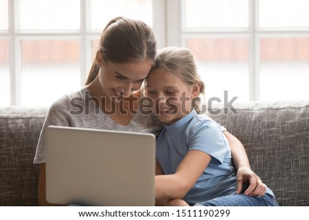 Happy young mother and teen daughter rest cuddling on sofa at home watching movie on laptop together, loving mom and girl child enjoy spending weekend relaxing browsing internet on computer