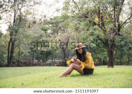 Beautiful Asian Bohemian girl with boho style dress and hat play social media, shop online, and chat by smartphone in park. Attractive female hipster rest in garden.