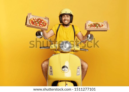 Photo of busy food courier drives fast motorbike, carries two carton boxes with delicious pizza, wears yellow helmet and casual t shirt, clenches teeth, being in hurry to bring order in time.