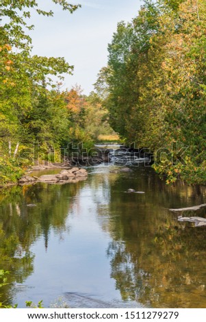 Scenic early fall landscape picture featuring creek with calm running water, trees, blue sky and tree reflections on the water