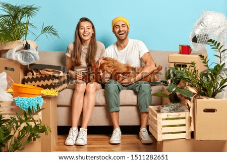 Positive delighted woman and man play with their favourite dog, pose on sofa, rejoice day of moving in new apartment, surrounded with different boxes, indoor plants. Family, relocation and fun concept Royalty-Free Stock Photo #1511282651