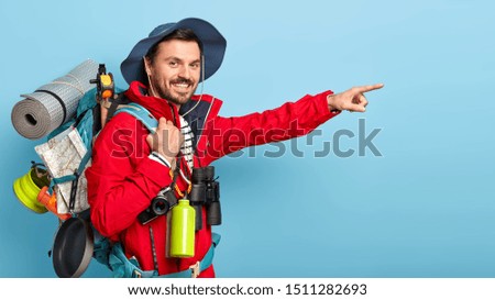 Profile shot of handsome cheerful man traveler points into distance, wears hat and red jacket, carries backpack with map, rolled up karemat, uses binoculars, isolated on blue wall, hikes in mountains