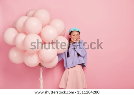 Overjoyed teenage girl keeps eyes closed, smiles broadly, shows white teeth, wears beret, sweatshirt and skirt, holds inflated balloons, celebrates getting bachelors degree, isolated on rosy wall