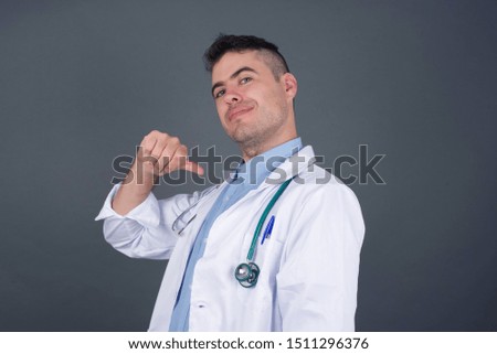 Closeup of cheerful European  young doctor man looks joyful, satisfied and confident, points at himself with thumb, isolated over gray studio background.