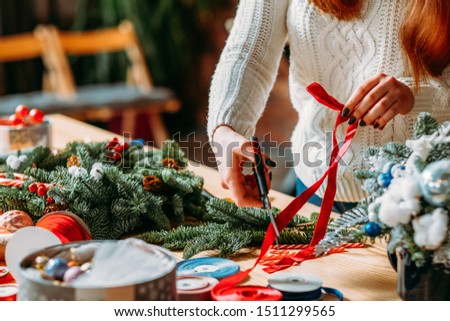 Craft workshop. Cropped shot of female florist using red ribbon, green fir tree twig to create Christmas interior decoration.