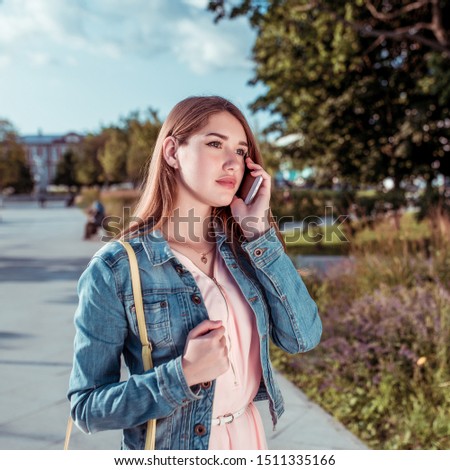 Girl in summer in city park, calling on her cell phone, in pink blouse and denim jacket, long hair, casual makeup. Emotions of comfort and attentively listens to interlocutor.