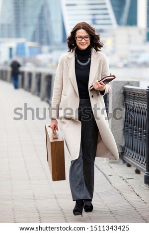A beautiful smiling woman in a light coat and with a wooden case in her hands walks along the autumn promenade against the background of skyscrapers. Fashion, style, business