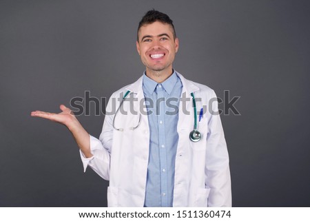 Positive glad doctor male says: wow how exciting it is, has amazed expression, shows something on blank space with open hand over isolated gray background. Advertisement concept.
