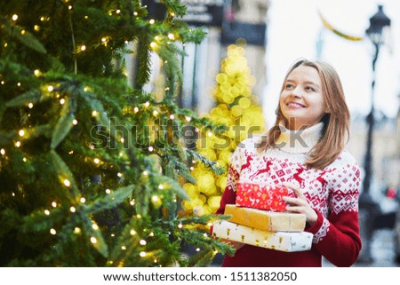 Happy young girl in warm red knitted sweater with pile of holiday gifts on a street of Paris decorated for Christmas