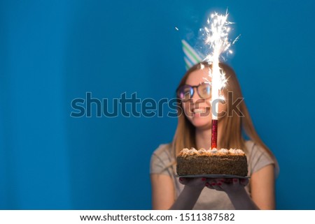 Cheerful young blurred girl student in glasses holding a congratulatory cake with a candle standing on a blue background. Birthday concept.