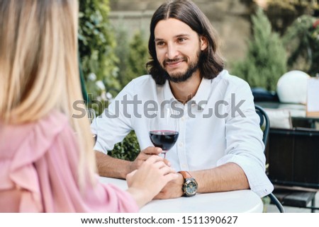 Young handsome brunette bearded man in shirt with glass of wine happily looking at girlfriend on romantic date in cafe outdoor