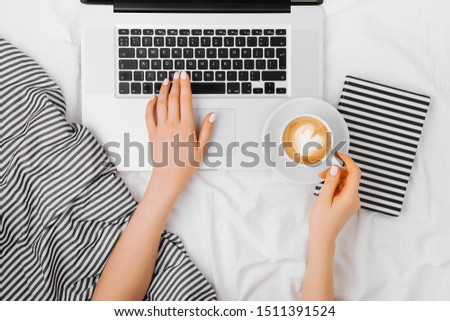 Young woman drinking coffee at home in her bed and working her laptop, top view