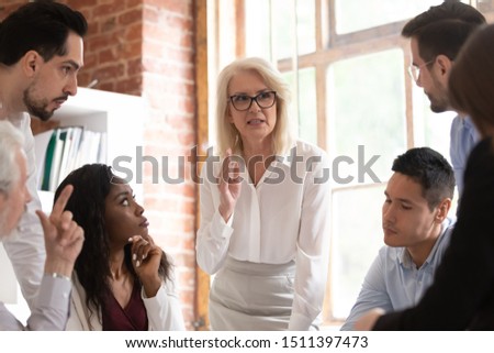 Middle aged businesswoman boss team leader defines tasks and corporate goals aims for multinational employees staff at meeting in office, mentoring brainstorming lead by older company member concept Royalty-Free Stock Photo #1511397473
