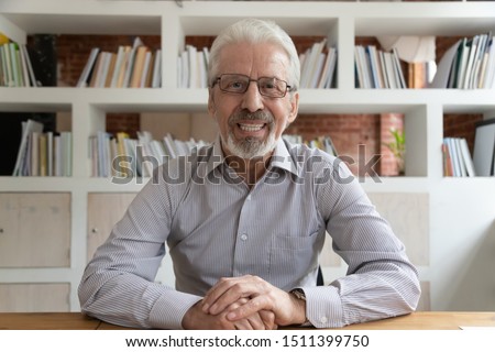 Positive aged grey-haired businessman in glasses sit at desk looking at camera, webcam view. Portrait of skilled financial advisor lead online course old entrepreneur and virtual communication concept Royalty-Free Stock Photo #1511399750