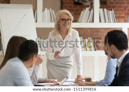Staff sit at boardroom desk listening middle-aged businesswoman company boss explaining current plans and goals, successful speaker tell about sales to investors clients or coach teach interns concept Royalty-Free Stock Photo #1511399816
