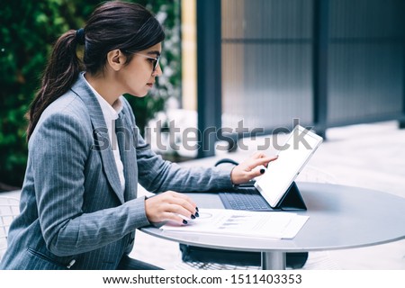 Side view of formally dressed female entrepreneur searching mistakes on online document report during correcting mistakes on paper report, Spanish woman working on touch pad with copy space for text
