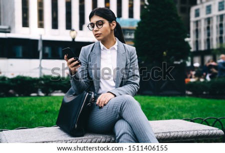 Spanish female entrepreneur in classic eyewear checking information on website on modern cellular gadget, executive businesswoman phoning and writing email message for send via mobile application